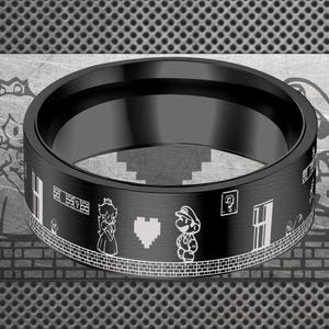 Puede incluir: Anillo negro con un diseño grabado en blanco que presenta personajes y elementos de un videojuego. El anillo tiene un acabado mate y una banda ancha. El diseño incluye una princesa, un fontanero, un corazón y otros gráficos relacionados con el juego.
