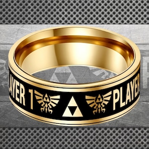 May include: A gold-colored ring with a black band featuring the text "PLAYER 1" and a video game logo. The ring has a polished finish and a smooth interior.