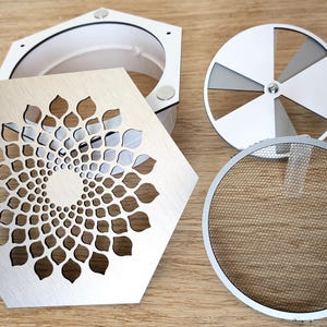 Op de afbeelding: Een assortiment metalen en houten componenten, waaronder een zeshoekig stuk met een bloemvormig uitgesneden ontwerp, een witte container, een cirkelvormig gaasscherm en een ventilatorachtige structuur. De items zijn gerangschikt op een houten oppervlak.