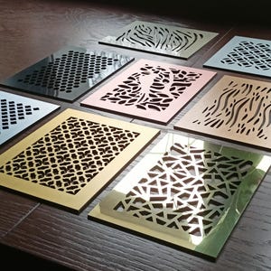 Op de afbeelding: Een verzameling decoratieve metalen ventilatieroosters met diverse uitgesneden patronen. De roosters zijn in verschillende kleuren, waaronder zwart, goud en zilver, en zijn gerangschikt op een houten oppervlak. De patronen omvatten geometrische vormen, organische ontwerpen en abstracte vormen.