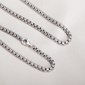 May include: A silver box chain necklace with a lobster clasp. The chain is composed of small, square-shaped links. The necklace is displayed on a white, textured surface.