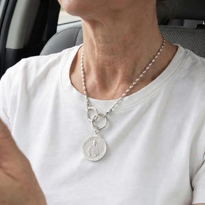 May include: A silver-toned necklace featuring a chain of small, oval-shaped beads. The chain connects to two interlocking circular rings, from which a round, silver-toned pendant hangs. The pendant displays a relief image of a figure in robes.