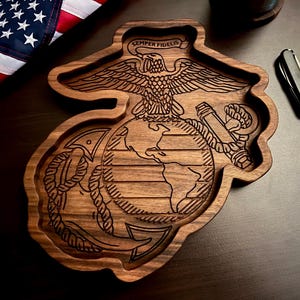 May include: A dark brown wooden tray shaped like the United States Marine Corps emblem, featuring an eagle, globe, and anchor design with the text Semper Fidelis engraved at the top. The tray has a smooth finish and follows the contour of the logo.