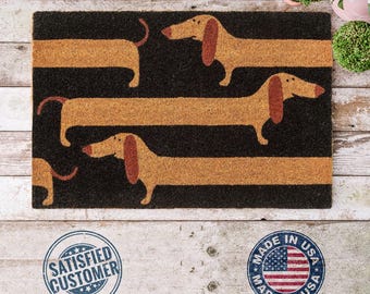 May include: Rectangular doormat with a black background and horizontal tan stripes. Tan dachshund dogs are printed on the tan stripes. The doormat has a "Satisfied Customer" stamp and a "Made in USA" stamp.