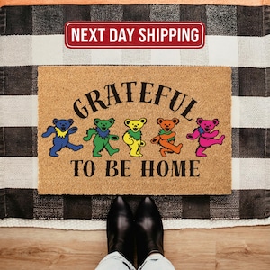 May include: A rectangular doormat with the words "Grateful To Be Home" and colorful dancing bear illustrations. The mat is made of a natural fiber material and is placed on a black and white checkered rug.