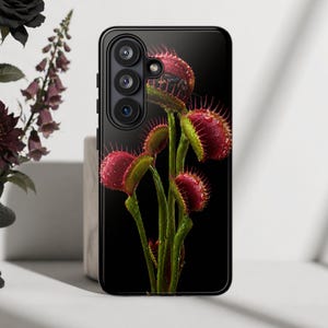 Puede incluir: Una funda de smartphone negra con un gráfico de alta resolución de varias plantas atrapamoscas sobre un fondo negro sólido. Las plantas tienen trampas rojas dentadas y tallos verdes, todo cubierto con pequeñas gotas de agua brillantes.