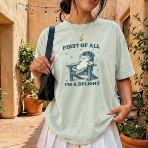 Op de afbeelding: Lichtgroen T-shirt met de tekst "FIRST OF ALL I'M A DELIGHT" en een afbeelding van een kikker in een stoel. Het shirt is in de kleur baai.