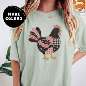 Puede incluir: Una camiseta de manga corta en color verde salvia claro con un gráfico central de una gallina compuesta por varios parches estampados en negro, blanco y rosa, incluyendo cuadros vichy y lunares. Un distintivo negro indica MORE COLORS.
