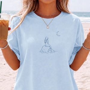 Puede incluir: Camiseta azul claro con un diseño minimalista de sirena y luna creciente en azul marino. El texto "CHAMBRAY COLOR" está impreso en la parte inferior. La camiseta tiene mangas cortas y un corte holgado.
