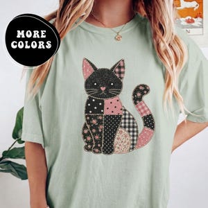 Puede incluir: Una camiseta verde claro con un gráfico de un gato sentado compuesto por varios patrones de patchwork en negro, blanco y rosa. El diseño del gato incluye texturas de tela con flores, lunares, estrellas y cuadros. El texto MORE COLORS es visible.
