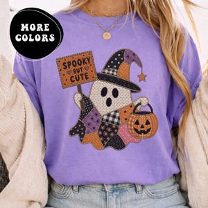 May include: A violet t-shirt featuring a graphic of a ghost wearing a patchwork witch hat and holding a pumpkin bucket. The ghost holds a sign that reads SPOOKY BUT CUTE. The design incorporates orange, black, and purple patterned fabric textures.