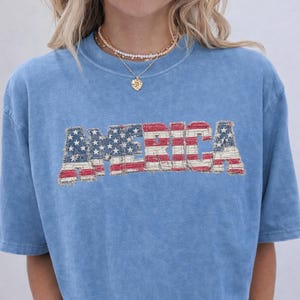 Puede incluir: Camiseta azul claro con la palabra "AMERICA" en un diseño desgastado inspirado en la bandera. El texto presenta rayas rojas y blancas y estrellas blancas sobre un fondo azul. La camiseta es de color vaquero azul.