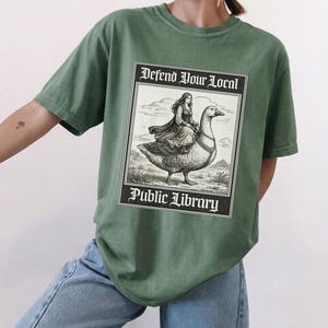 Puede incluir: Camiseta verde con un gráfico que dice "Defend Your Local Public Library". El gráfico representa a una mujer montada en un ganso. La camiseta es de color verde liso.