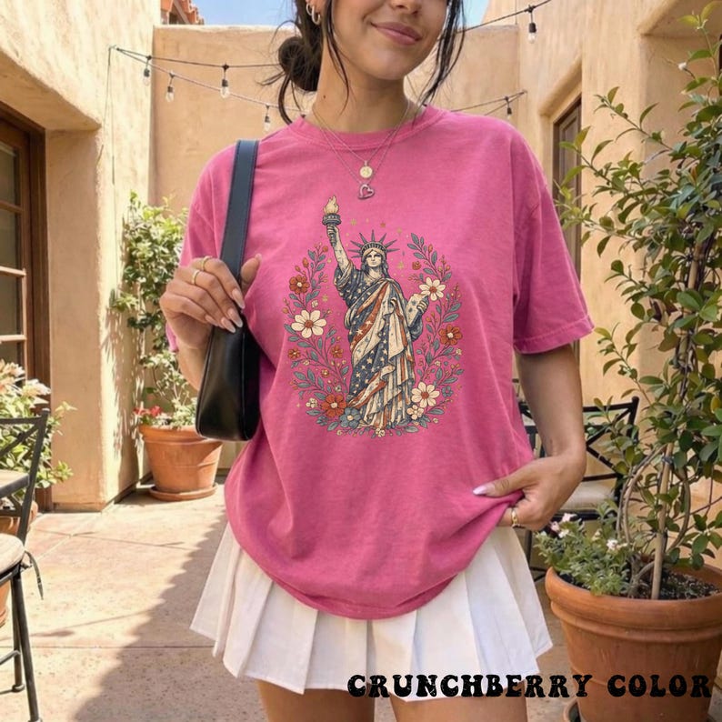 May include: A pink t-shirt featuring a graphic of the Statue of Liberty surrounded by flowers. The statue is depicted in red, white, and blue. The text "CRUNCHBERRY COLOR" is printed at the bottom of the shirt.