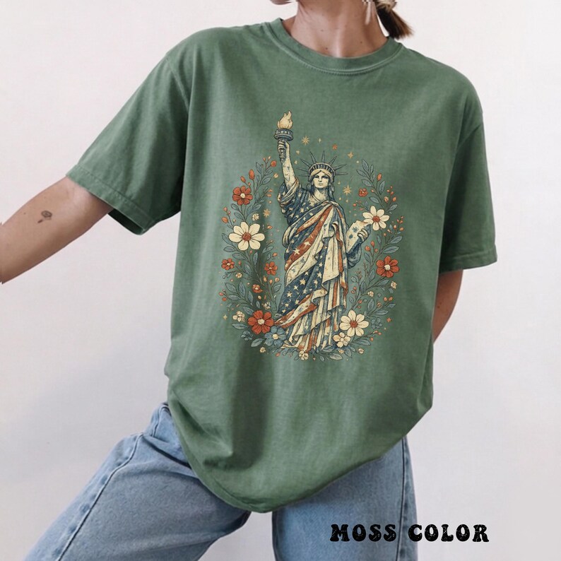 May include: Moss green t-shirt featuring a graphic of the Statue of Liberty surrounded by a floral wreath. The Statue of Liberty is depicted in red, white, and blue, holding a torch. The text "MOSS COLOR" is at the bottom of the shirt.
