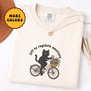 Puede incluir: Camiseta color crema con un gato negro montando en bicicleta, llevando una taza de café y flores. El texto "Off to regulate emotions" está impreso en la camiseta. Un collar dorado está sobre la camiseta.