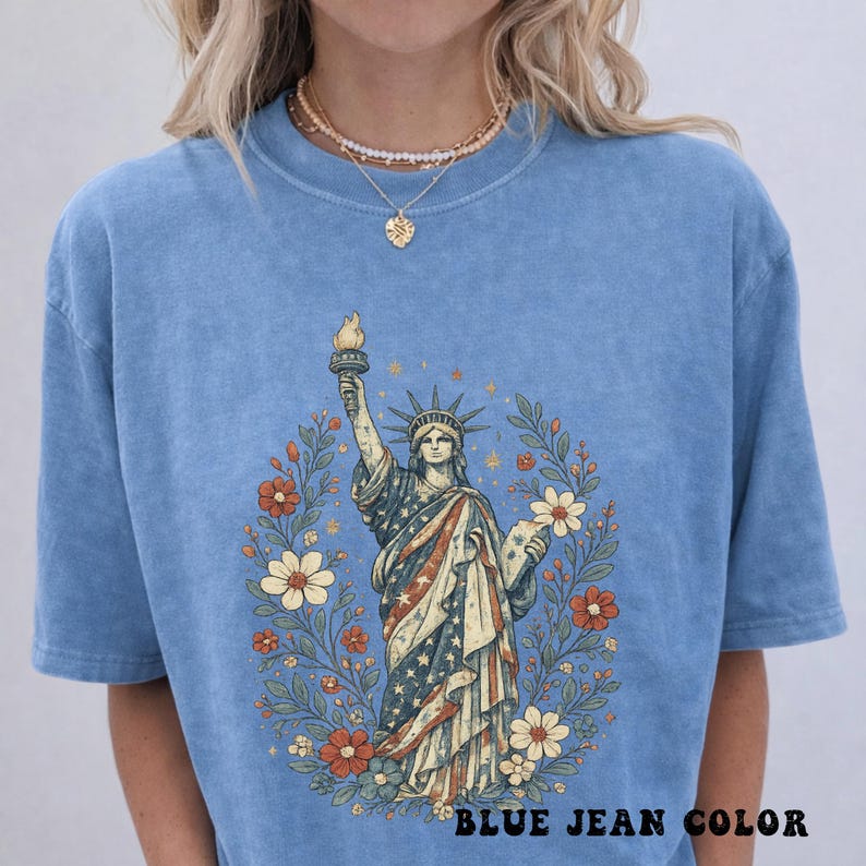 May include: Blue jean-colored t-shirt featuring a graphic of the Statue of Liberty, adorned with a floral wreath. The statue is draped in the American flag's colors. The text "BLUE JEAN COLOR" is at the bottom.