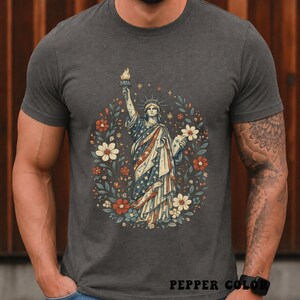 May include: Dark gray t-shirt featuring a graphic of the Statue of Liberty in a floral wreath. The statue is draped in a flag-themed design with stars and stripes. The shirt has the text "PEPPER COLOR" at the bottom.