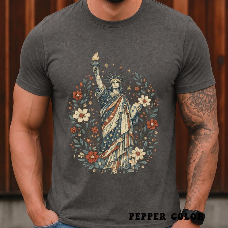 May include: Dark gray t-shirt featuring a graphic of the Statue of Liberty in a floral wreath. The statue is draped in a flag-themed design with stars and stripes. The shirt has the text "PEPPER COLOR" at the bottom.
