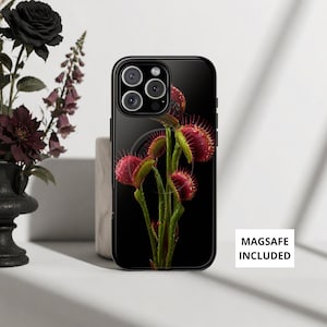 Puede incluir: Una funda de smartphone negra con un gráfico de plantas atrapamoscas verdes y rojas sobre un fondo negro. La funda tiene un diseño de anillo MagSafe circular visible en la parte posterior. El texto MAGSAFE INCLUDED está impreso en el lateral.