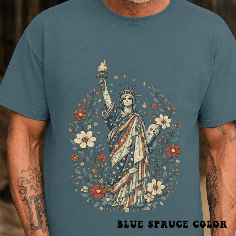 May include: Blue spruce color t-shirt featuring a detailed illustration of the Statue of Liberty holding a torch, surrounded by a floral wreath. The statue's robe is adorned with stars and stripes. The text "BLUE SPRUCE COLOR" is at the bottom.