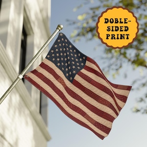 May include: A fabric American flag featuring a dark blue canton with white stars and alternating red and cream-colored stripes with a quilted texture. The flag is attached to a metal pole and includes a yellow circular badge stating DOBLE-SIDED PRINT.