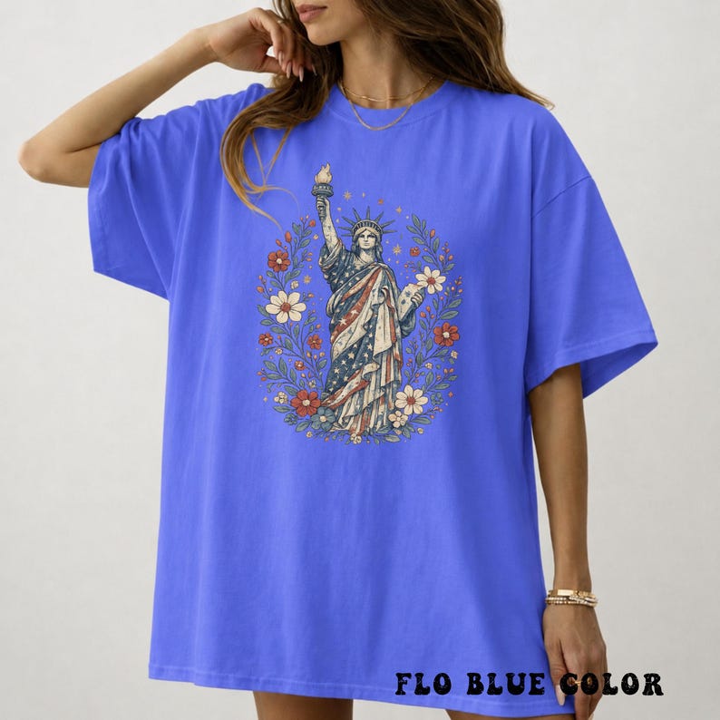 May include: A blue t-shirt featuring a graphic of the Statue of Liberty surrounded by a floral wreath. The statue is depicted in red, white, and blue, holding a torch. The text "FLO BLUE COLOR" is printed at the bottom of the shirt.