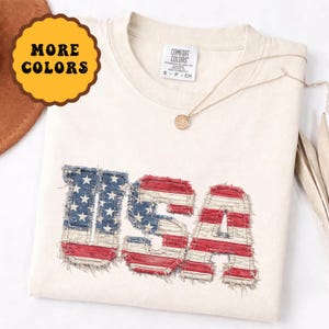 Puede incluir: Camiseta color crema con la palabra "USA" en un diseño desgastado con los colores de la bandera estadounidense. Las letras parecen estar hechas de mezclilla y rayas rojas y blancas. También se ven un collar dorado y un botón "More Colors".
