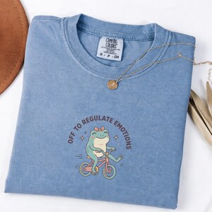 Op de afbeelding: Een lichtblauw Comfort Colors T-shirt met de tekst "OFF TO REGULATE EMOTIONS" en een cartoon kikker op een fiets. Een gouden ketting met een hanger ligt op het shirt. Het shirt heeft een klein label met "S - P - CH".