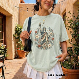 May include: A light green t-shirt featuring a graphic of the Statue of Liberty surrounded by flowers. The statue is depicted in red, white, and blue, with a floral wreath. The text "BAY COLOR" is at the bottom of the shirt.