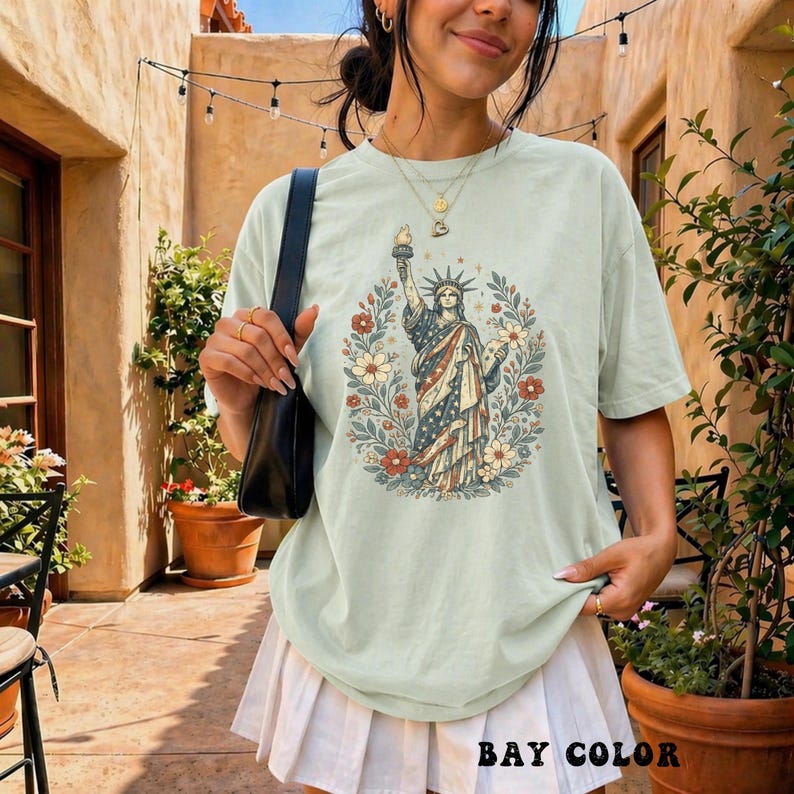 May include: A light green t-shirt featuring a graphic of the Statue of Liberty surrounded by flowers. The statue is depicted in red, white, and blue, with a floral wreath. The text "BAY COLOR" is at the bottom of the shirt.