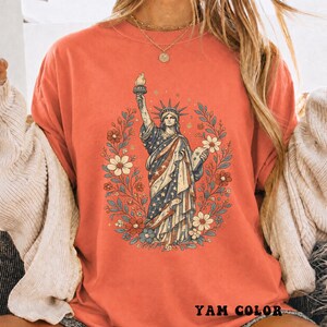 May include: Coral-colored t-shirt featuring a graphic of the Statue of Liberty surrounded by a floral wreath. The statue is depicted with the American flag's stars and stripes. The text "YAM COLOR" is printed at the bottom.