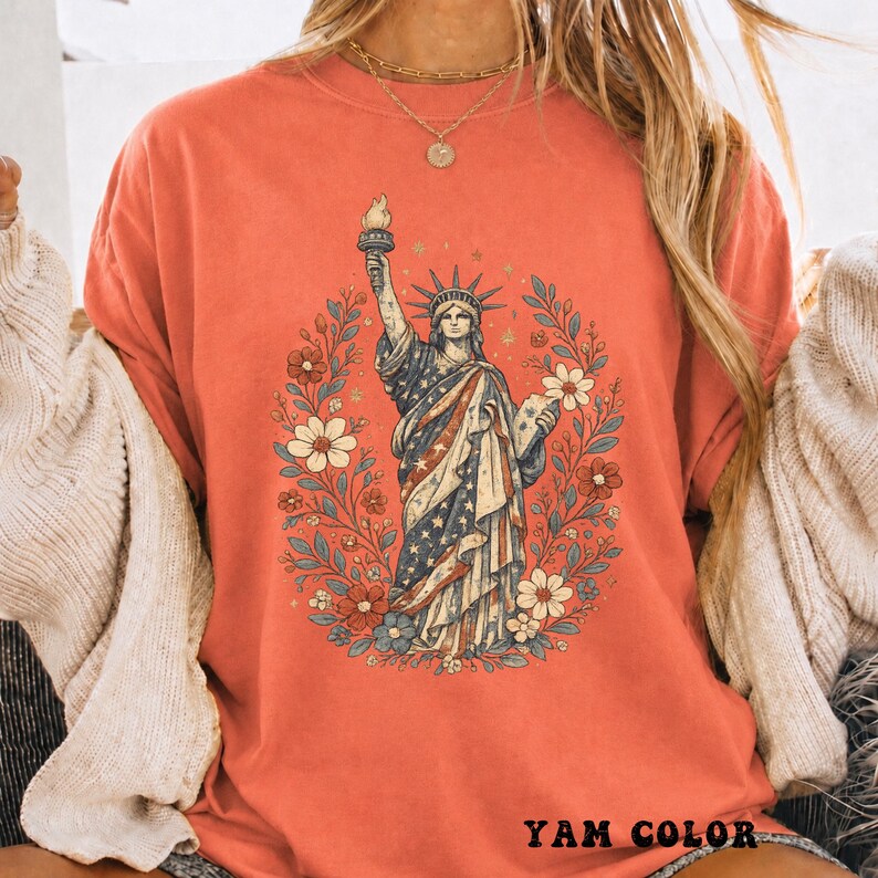 May include: Coral-colored t-shirt featuring a graphic of the Statue of Liberty surrounded by a floral wreath. The statue is depicted with the American flag's stars and stripes. The text "YAM COLOR" is printed at the bottom.
