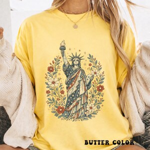 May include: Butter yellow t-shirt featuring a graphic of the Statue of Liberty with a floral wreath. The Statue of Liberty is depicted in red, white, and blue, holding a torch. The text "BUTTER COLOR" is printed at the bottom.