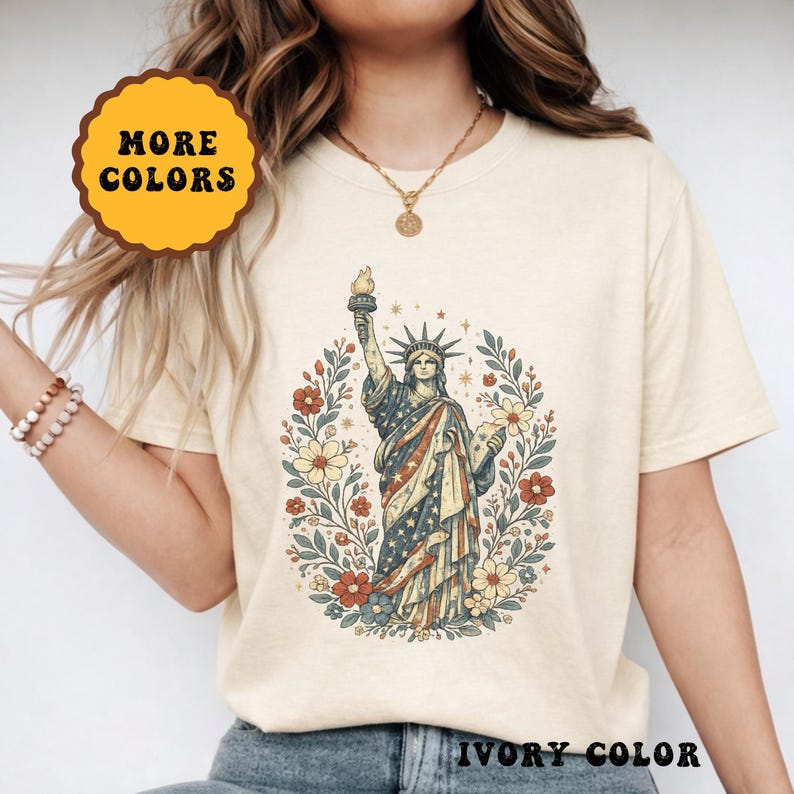 May include: Ivory-colored t-shirt featuring a graphic of the Statue of Liberty with a floral wreath. The statue is depicted in patriotic colors, holding a torch. The shirt has the text "MORE COLORS" and "IVORY COLOR".