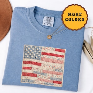 Puede incluir: Una camiseta azul claro con un diseño de bandera estadounidense cosido. La bandera presenta rayas rojas y blancas con estampados florales y estrellas de flores blancas. Un collar con colgante dorado descansa sobre la camisa. La etiqueta dice "Comfort Colors".
