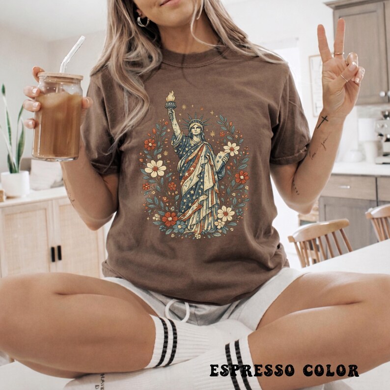 May include: Espresso-colored t-shirt featuring a graphic of the Statue of Liberty surrounded by flowers. The statue is depicted in red, white, and blue, holding a torch. The text "ESPRESSO COLOR" is printed at the bottom of the image.