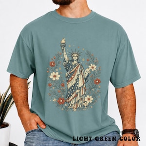 May include: Light green t-shirt featuring a graphic of the Statue of Liberty with a floral wreath. The Statue of Liberty is depicted in red, white, and blue, holding a torch. The text "LIGHT GREEN COLOR" is at the bottom of the shirt.