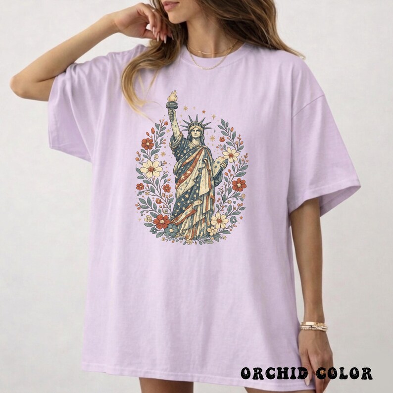 May include: A light purple t-shirt featuring a graphic of the Statue of Liberty in a floral wreath. The statue is depicted with the American flag colors. The text "ORCHID COLOR" is printed at the bottom of the shirt.
