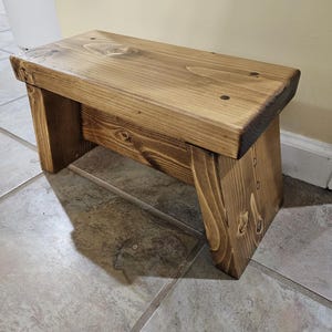 May include: A small, stained wooden step stool. The stool is rectangular with a flat top and angled legs. The wood has a warm, brown tone with visible grain and knots. The step stool is designed for use in the home.