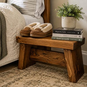 May include: A low, rectangular wooden bench with a dark brown stained finish and angled legs. On top sits a pair of brown suede slippers with cream-colored faux fur lining, a stack of three books, and a small potted green plant in a beige pot.