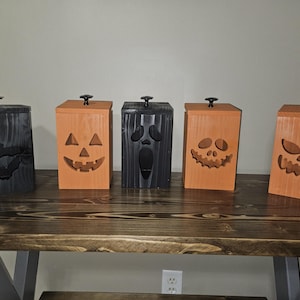May include: Five rectangular wooden boxes with lids and black knobs, arranged in a row. The boxes are painted in alternating black and orange colors, each featuring a unique cutout design including bats, a jack-o'-lantern, and ghost faces.