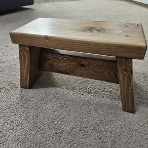 May include: A small, rectangular wooden step stool with a natural finish. The stool has a simple design with four legs and a flat top. The wood grain is visible, adding to its rustic charm. The stool measures approximately 12 inches long, 8 inches wide, and 6 inches high.