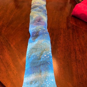 May include: A long, rectangular fabric item with a celestial pattern. The fabric features a blend of blues, purples, and white speckles, resembling a starry night sky. The item appears to be a decorative or functional textile, possibly a pillow or wrap.