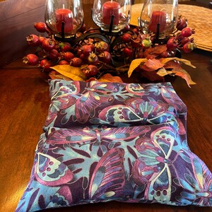 May include: A rectangular pillow with a butterfly pattern in shades of blue, purple, and teal. The pillow is resting on a wooden table. In the background, there are three glass candle holders with red candles and decorative berries.