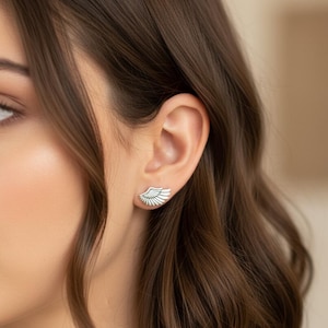 May include: A pair of silver-colored wing-shaped earrings. The earrings are worn on the ear of a person with brown hair. The earrings have a detailed feather design and are a stylish accessory.