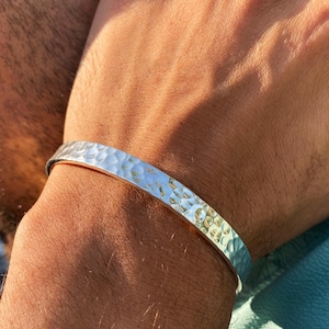 May include: A silver-toned cuff bracelet featuring a hammered, textured surface with multiple indentations. The metal band has a polished finish that reflects light across its uneven, dimpled exterior, creating a distinct pattern along the entire length.
