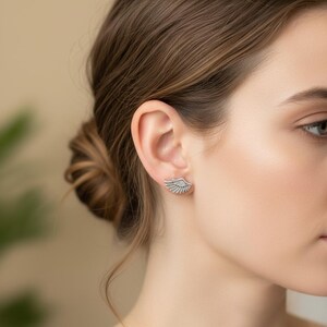 May include: Silver-toned wing-shaped stud earrings. The earrings are designed to be worn on the earlobe, with a detailed feather pattern. The earrings are shown on a person's ear, with a blurred background.