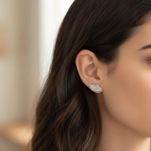 May include: A pair of silver-colored wing-shaped earrings. The earrings are worn on the ear and have a detailed feather design. The earrings are a jewelry item and are likely made of metal.