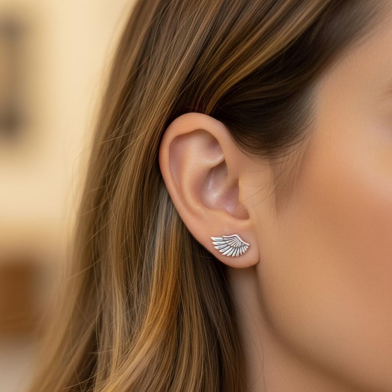 May include: Silver wing-shaped stud earrings. The earrings are worn on the ear, with the wing design facing outwards. The earrings are a bright silver color and have a detailed feather design. The earrings are small and delicate.